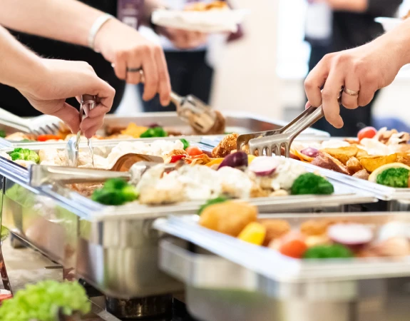 Catering dishes full of vegetables being served to people with tongs.