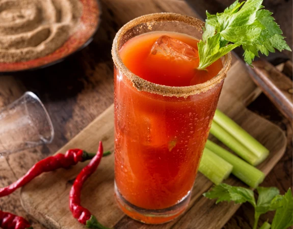 A caesar cocktail on a wooden cutting board with celery, peppers, and hummus in the background